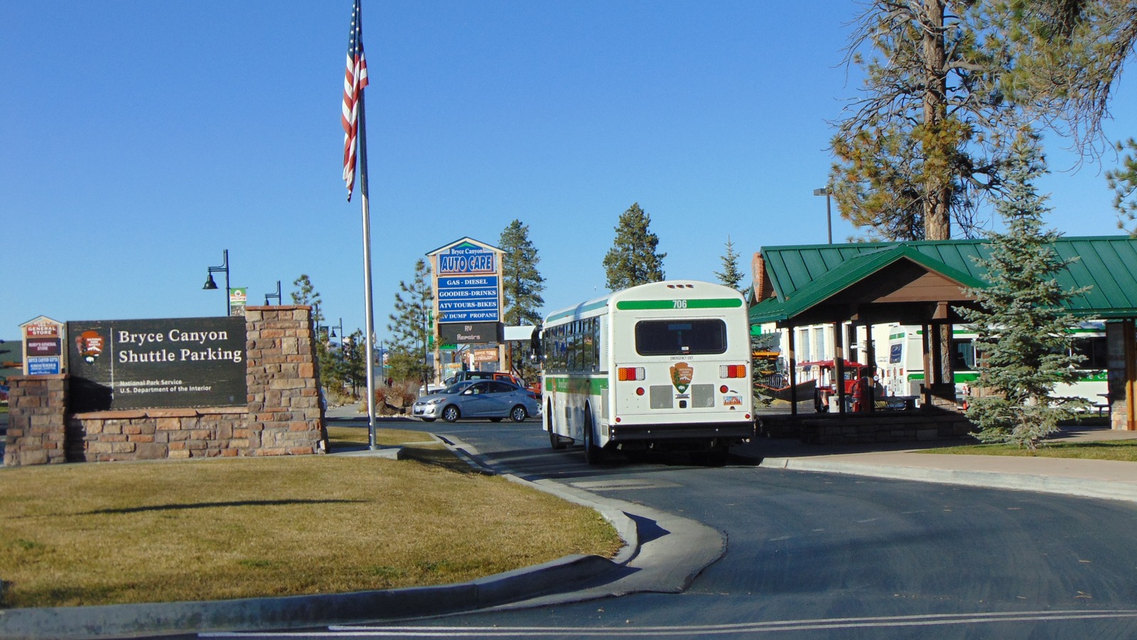 A white and green Bryce Canyon shuttle bus with a National Park Service arrowhead pulling away from the Bryce Canyon Shuttle Parking sign in Bryce Canyon City under a clear blue sky.