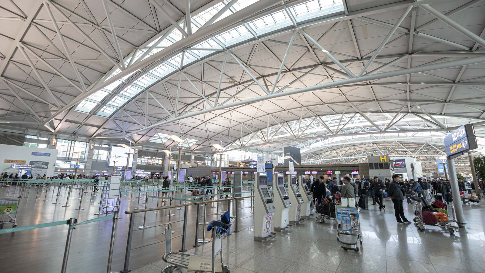 The departure hall of Terminal 1 at Incheon International Airport, its lattice roof over rows of check in desks and self service kiosks with passengers and luggage trolleys.