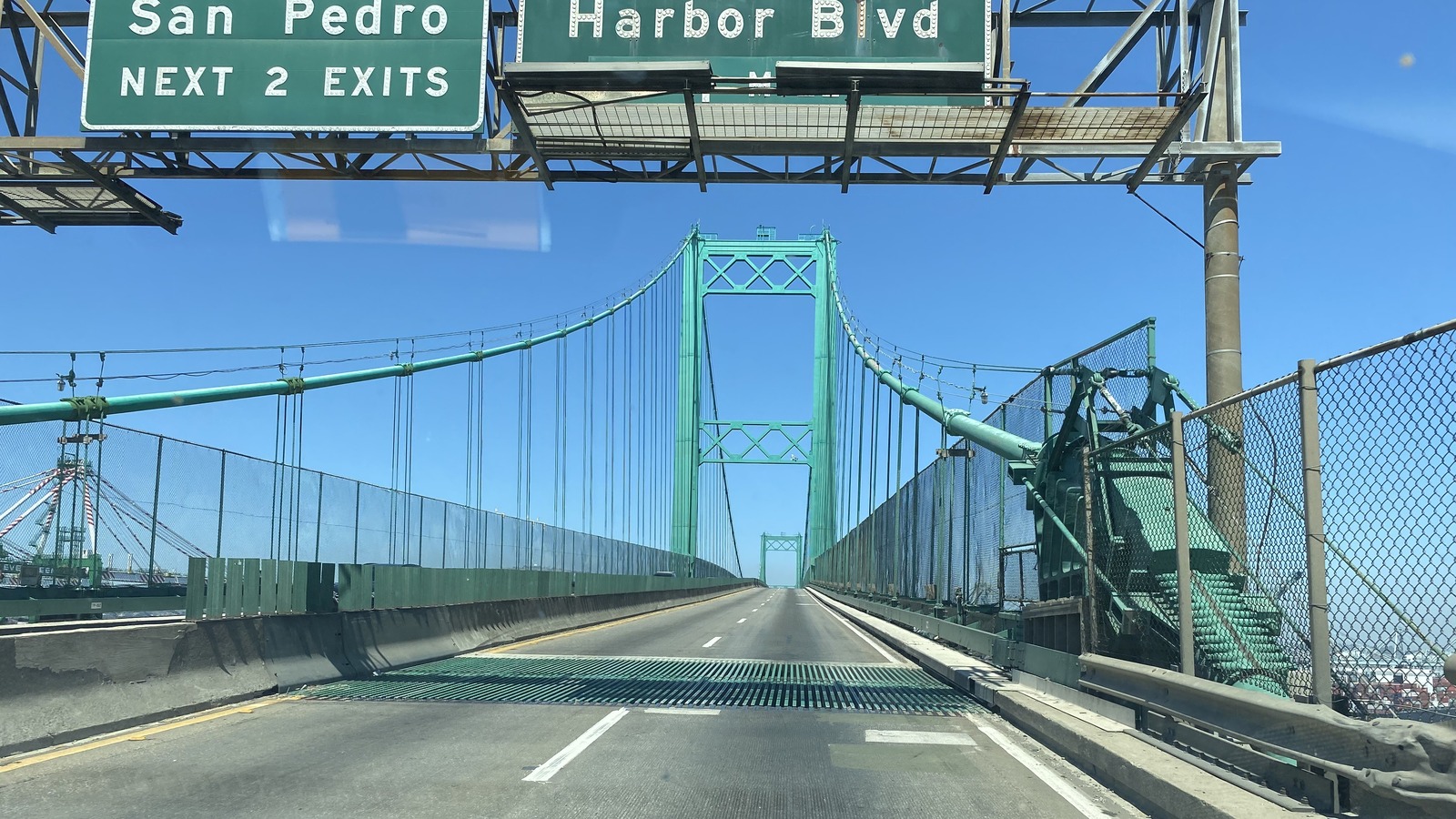 Driver view along the Vincent Thomas Bridge deck toward its green suspension towers, with San Pedro highway signs overhead