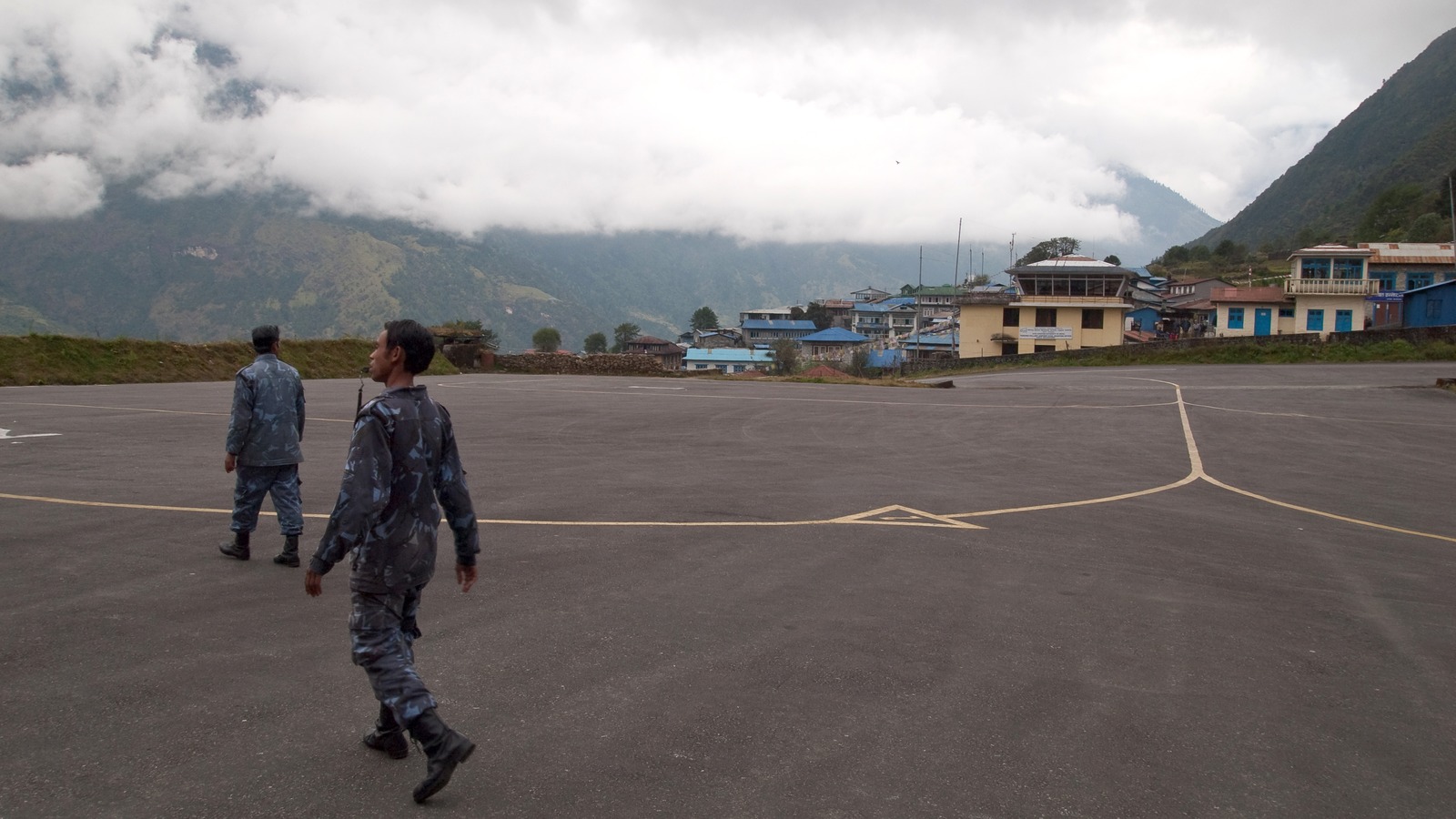 Tenzing-Hillary Airport apron and hillside terminal in Lukla, Nepal, under low cloud, with two people walking on the tarmac.