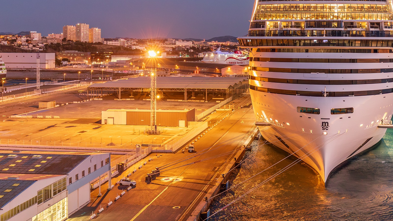 The bow of the MSC cruise ship MSC World Europa berthed at the Grand port maritime de Marseille at dusk, with the lit quay apron, terminal sheds and the Marseille hillside behind