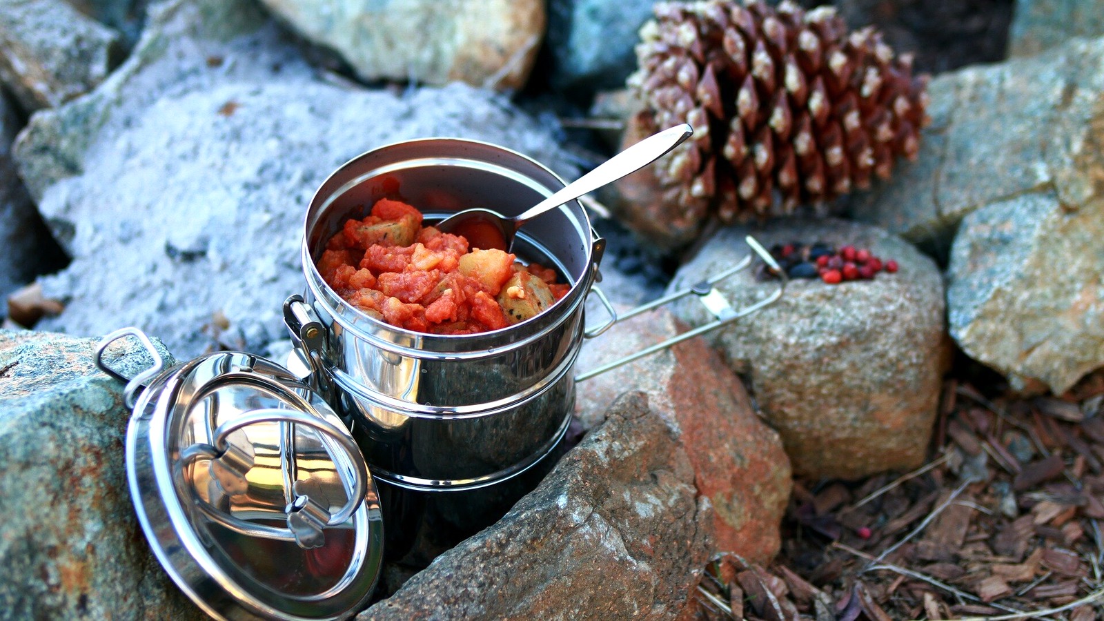 Stainless camping pot with lid and spoon resting among rocks outdoors.