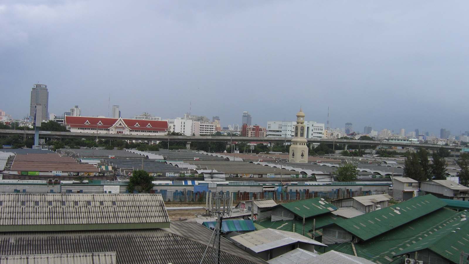 Rows of market roofs at Chatuchak Weekend Market in Bangkok stretch toward the city skyline under gray clouds.