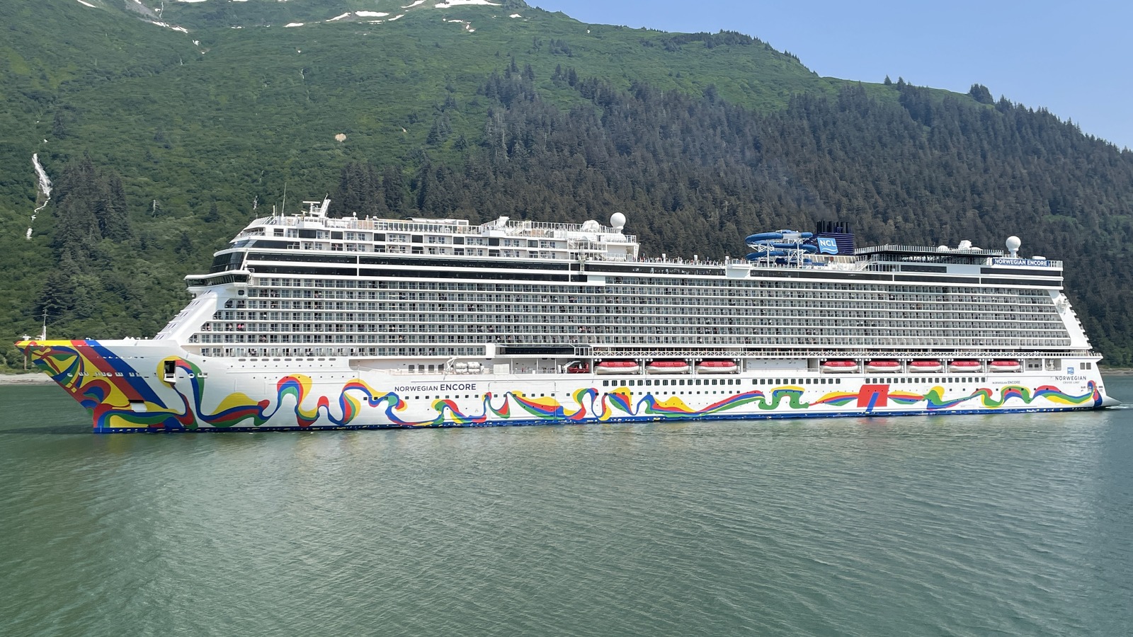 Norwegian Encore cruise ship on the water in Juneau, Alaska, with forested mountains behind the hull