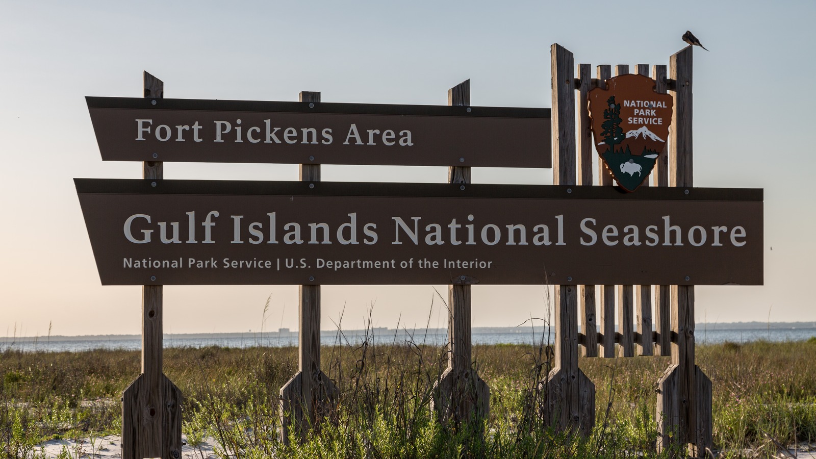 Fort Pickens Area entrance sign for Gulf Islands National Seashore with the National Park Service arrowhead, dunes, and water beyond