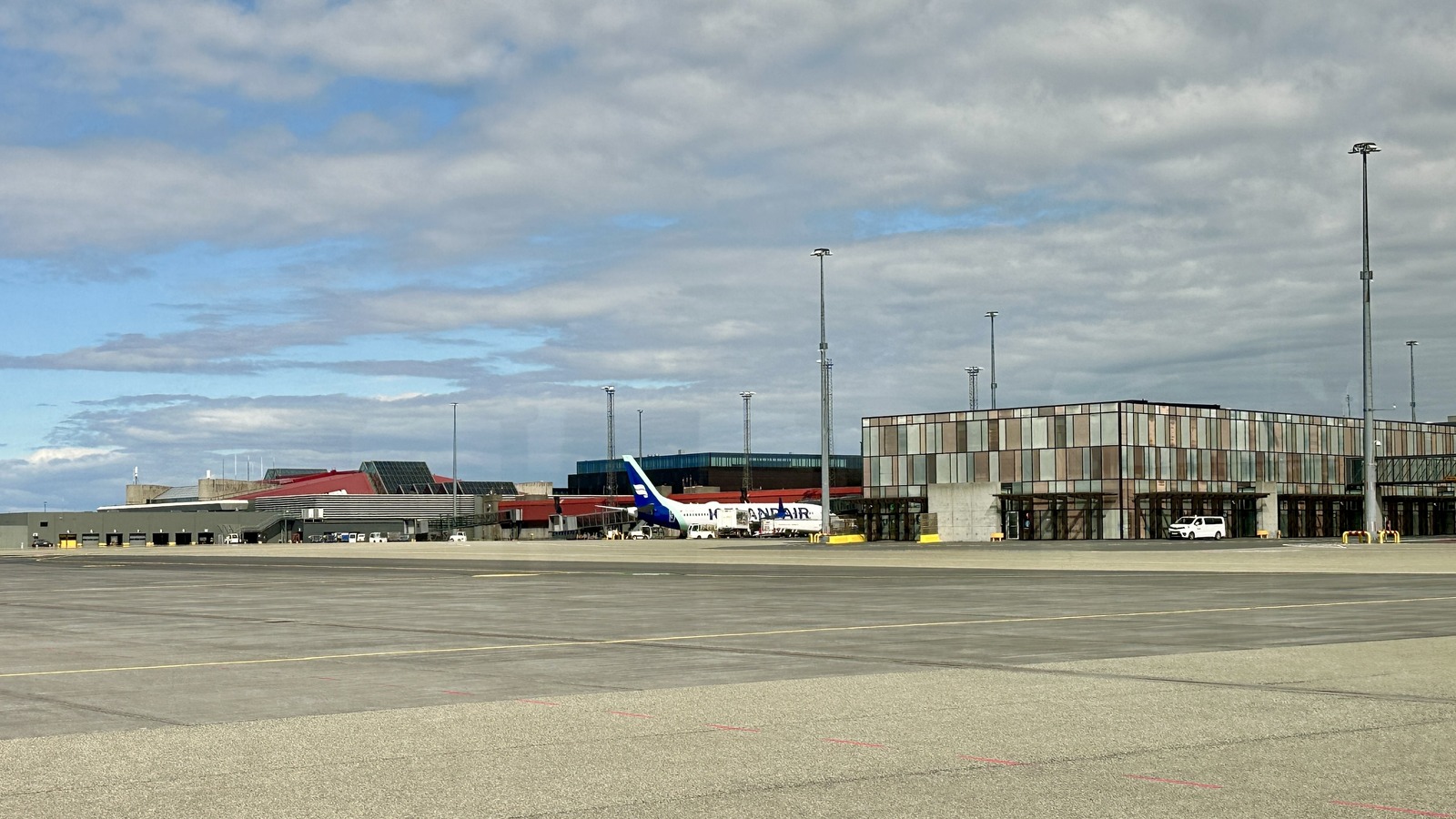 Keflavik International Airport terminal buildings seen across the apron under a partly cloudy sky, with an Icelandair jet parked at a gate.