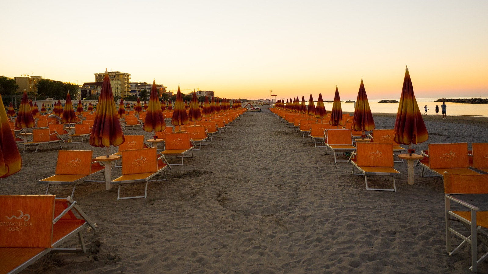Rows of orange beach umbrellas and lounge chairs on Rimini Beach at sunset, looking toward the Adriatic.