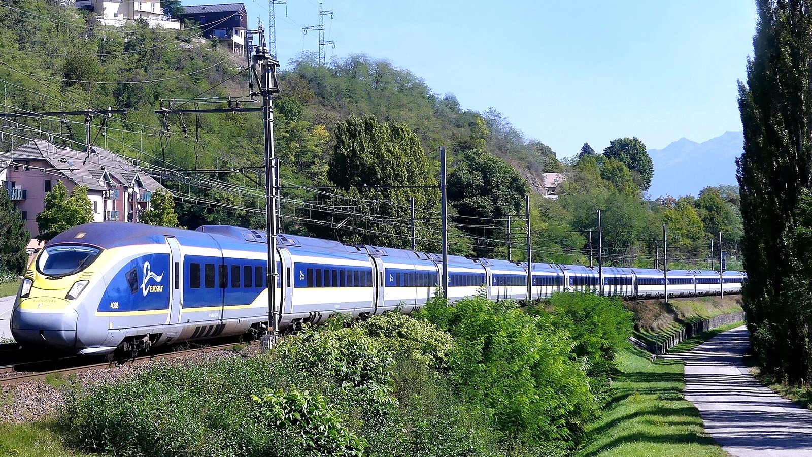 A blue and yellow Eurostar e320 high-speed train curving along an electrified line below a wooded hillside in Savoie, France