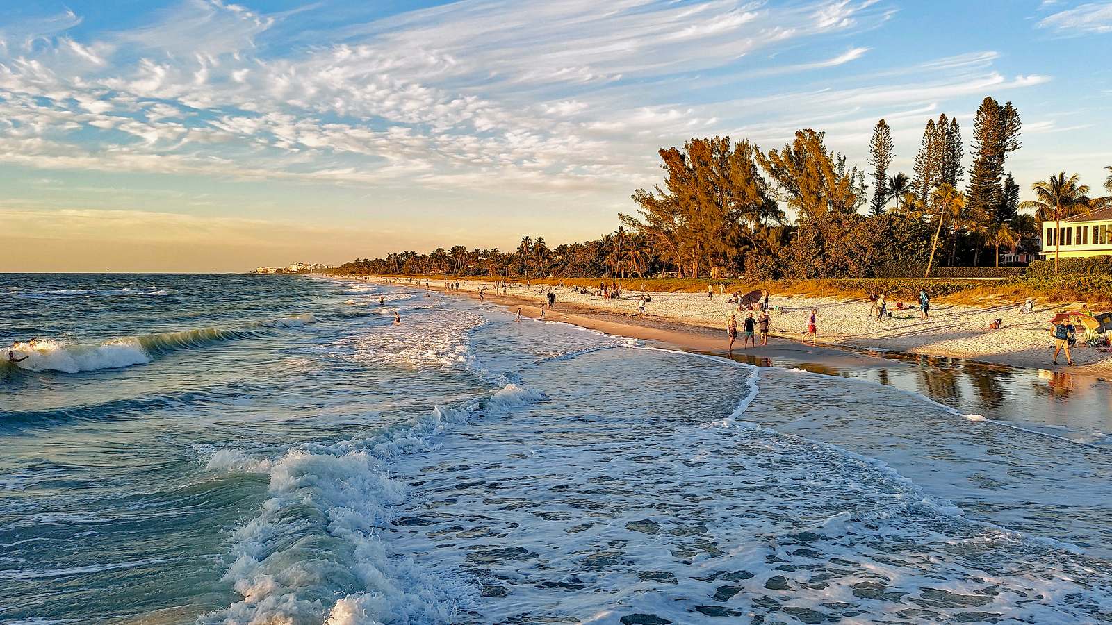 Waves wash onto Naples Beach on the Gulf at sunset, with palms and pines along the sand.