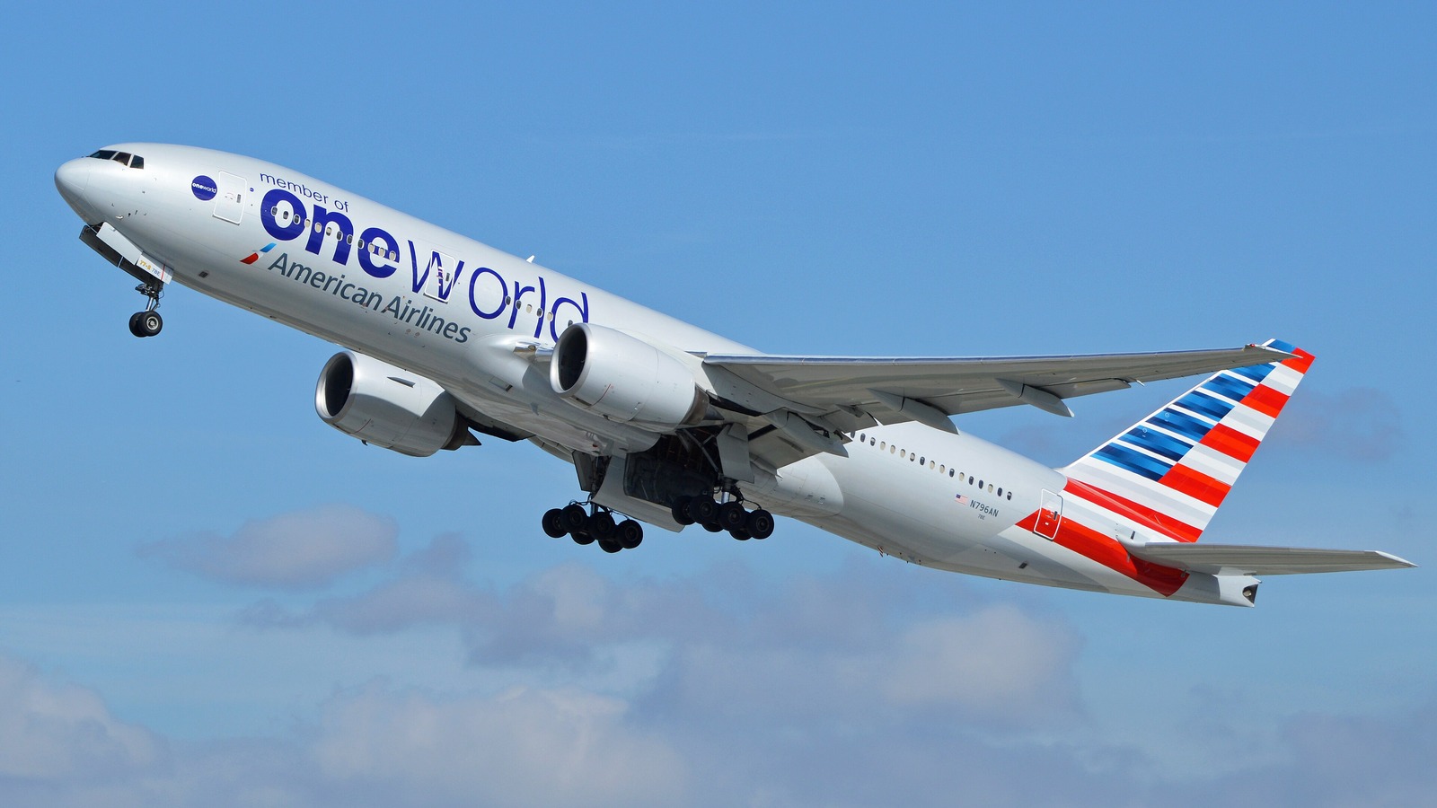 An American Airlines Boeing 777 painted in oneworld alliance livery climbing after takeoff against a blue sky.