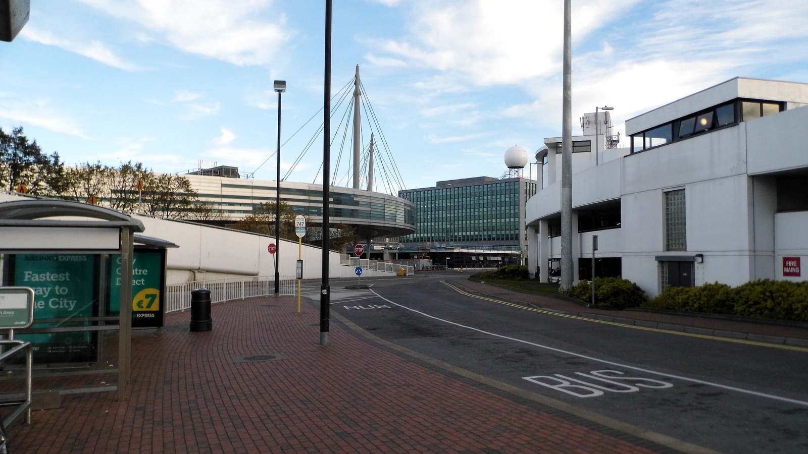 Landside road and buildings at Dublin Airport, including the circular control-tower complex and Airlink bus stop, under a bright sky.