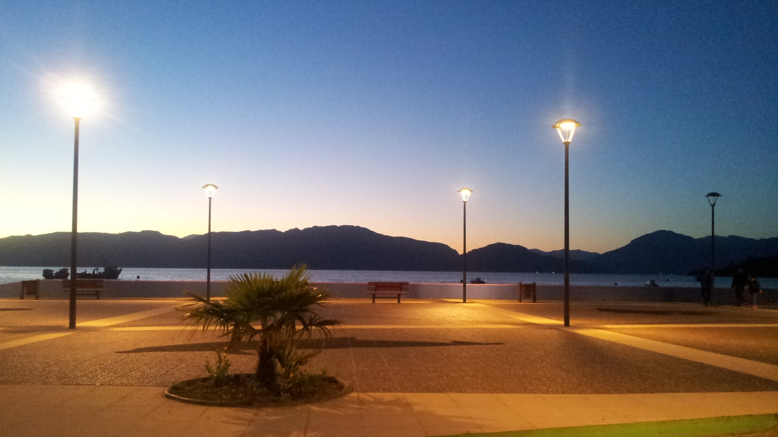 Waterfront promenade at Puerto Cisnes, Chile, at dusk with lamp posts, benches, and mountains across the channel