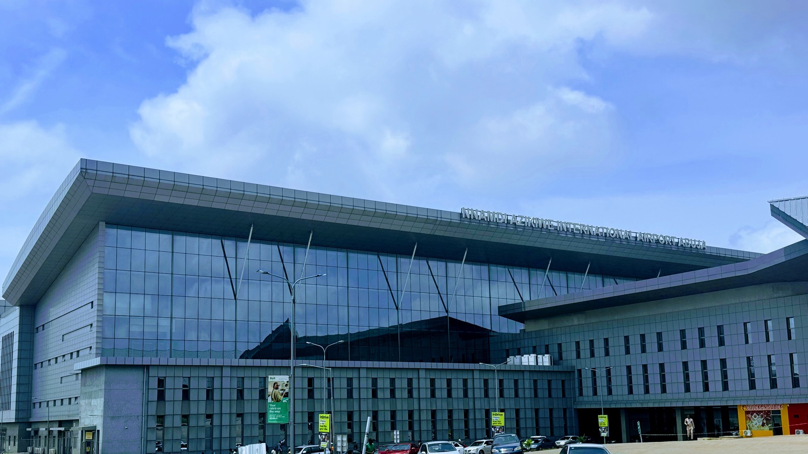 Exterior of the main terminal at Nnamdi Azikiwe International Airport in Abuja, with the airport name on the facade and vehicles at the drop-off.
