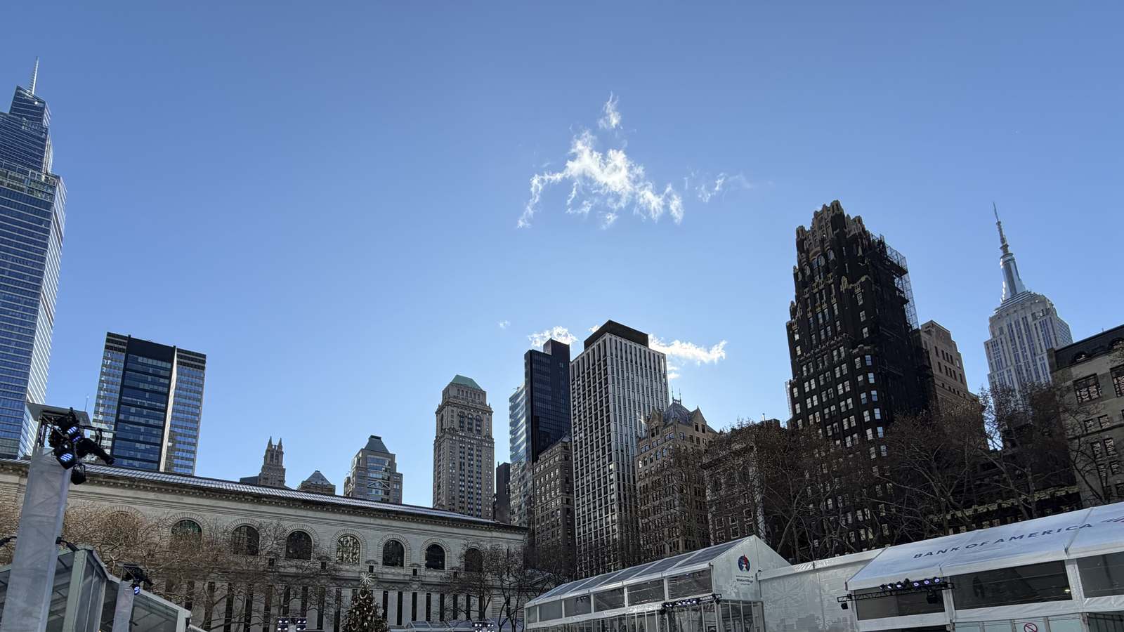 Bank of America Winter Village tents in Bryant Park under a blue sky, with the New York Public Library and the Empire State Building behind.