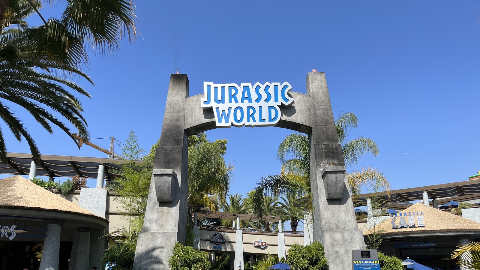 Guests walk under the Jurassic World entrance arch at Universal Studios Hollywood on a clear day