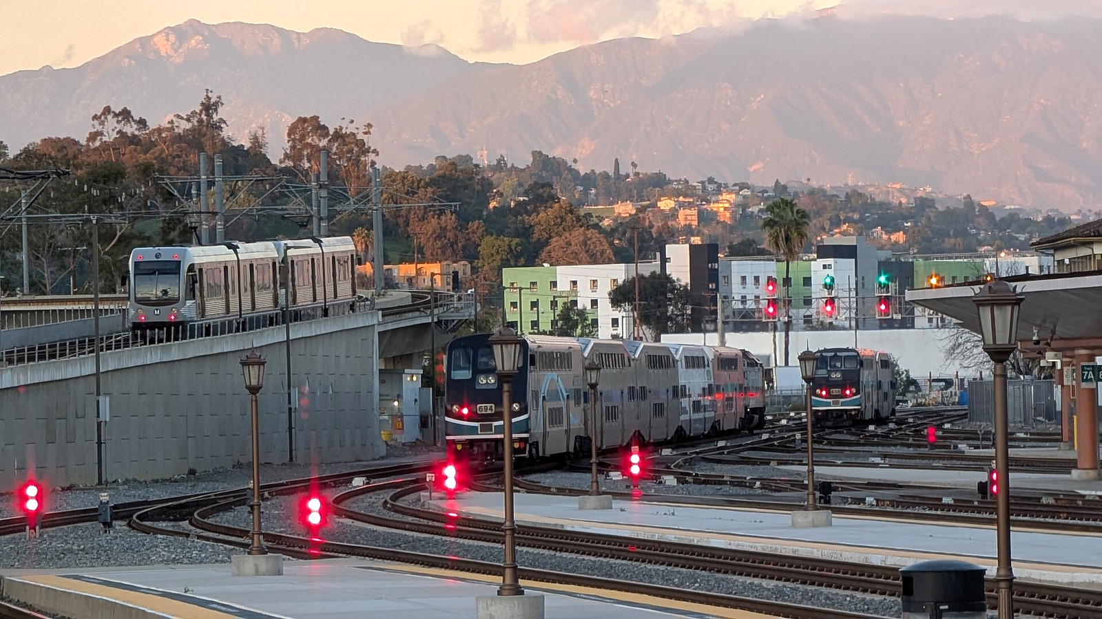 A Los Angeles Metro train and two Metrolink trains at Los Angeles Union Station near dusk with mountains behind