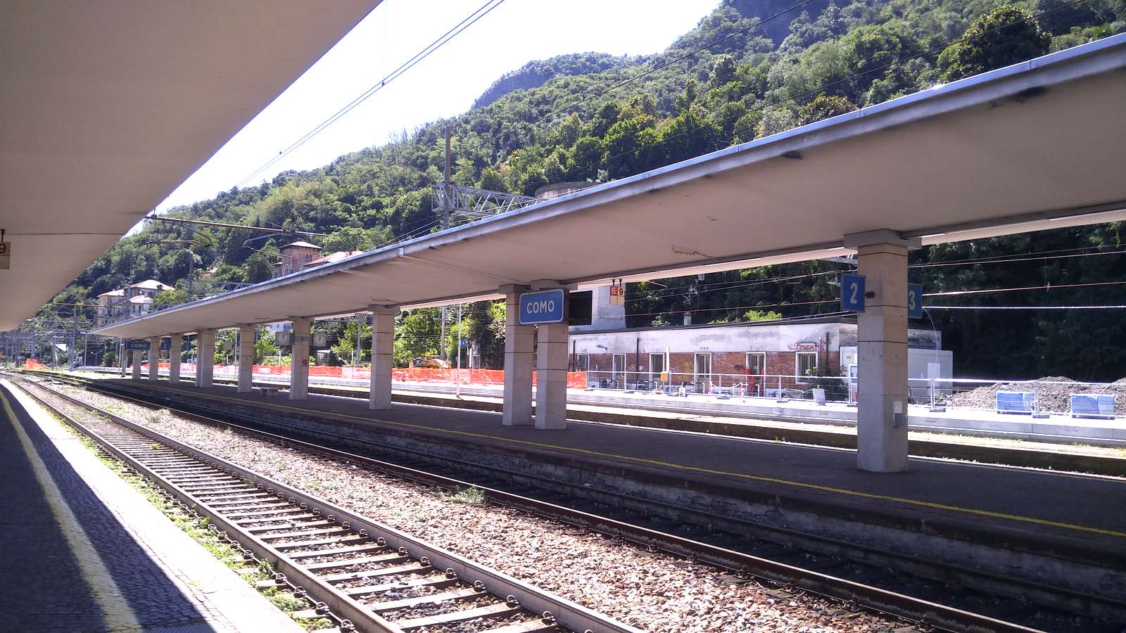 Platforms at Como San Giovanni station with the Como sign and wooded hillside behind