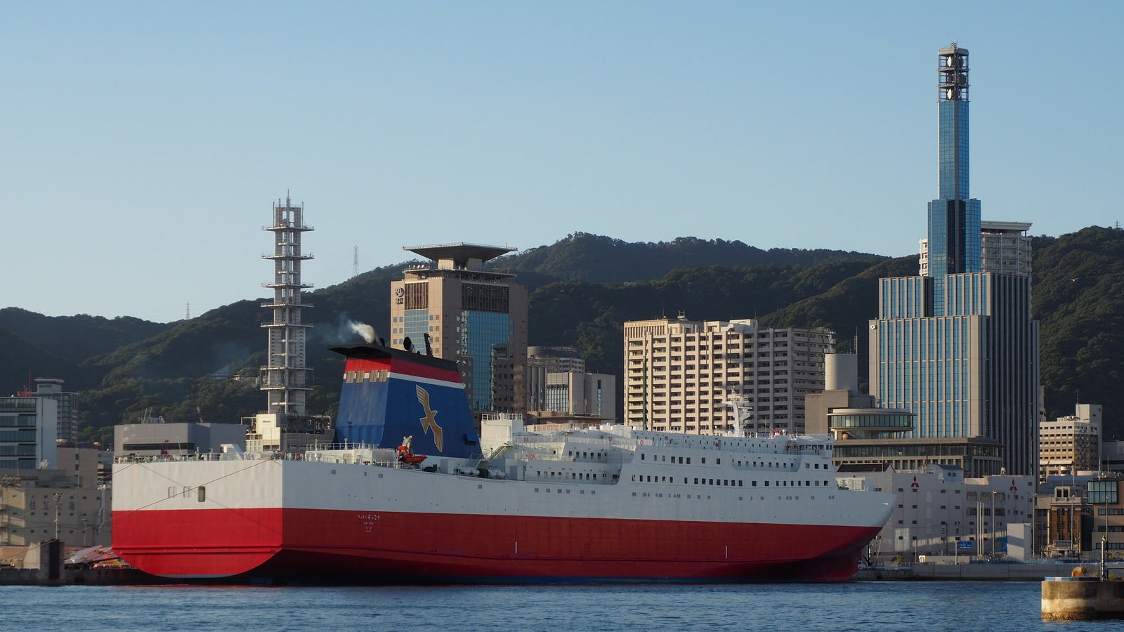 Miyazaki Car Ferry ship Ferry Rokko docked at the Port of Kobe, with a red hull, white superstructure, and blue funnel with a gold bird, against the Kobe waterfront.