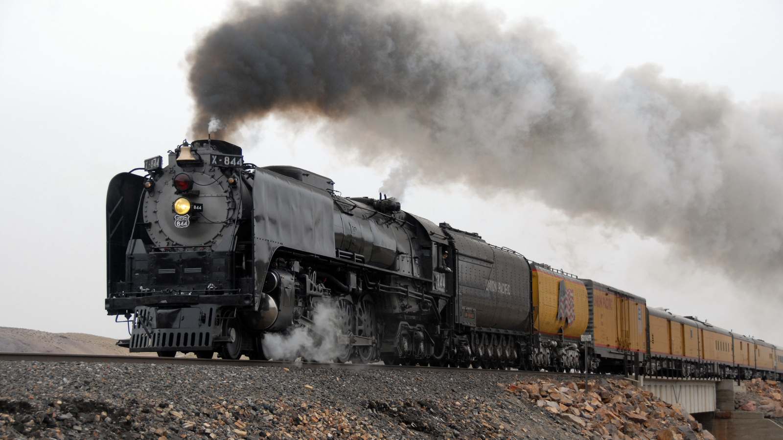 Union Pacific steam locomotive No. 844 pulls a line of yellow cars across a desert landscape under a plume of dark smoke.