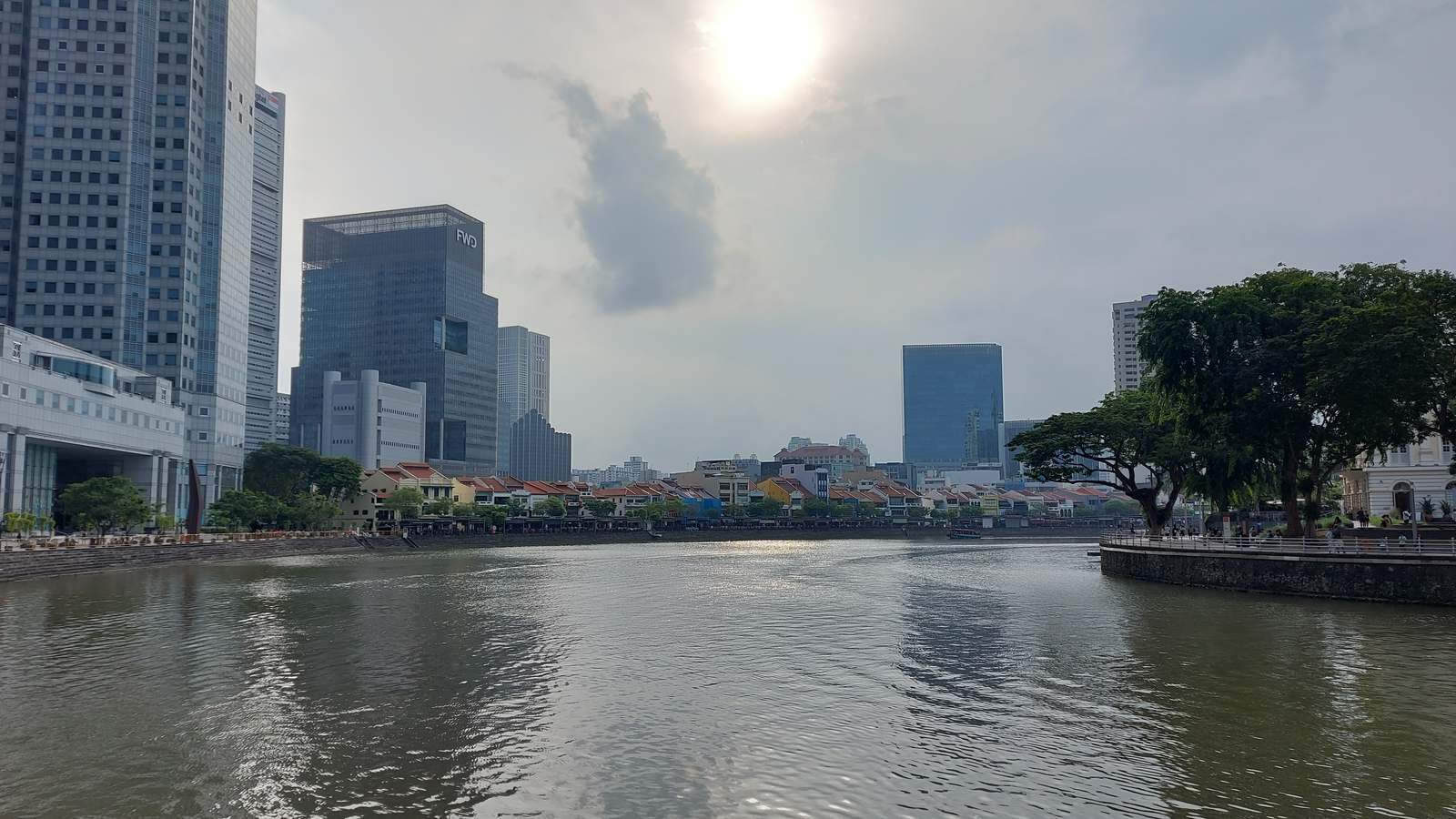 The Singapore River curving past low-rise shophouses and office towers in central Singapore under a hazy sky.
