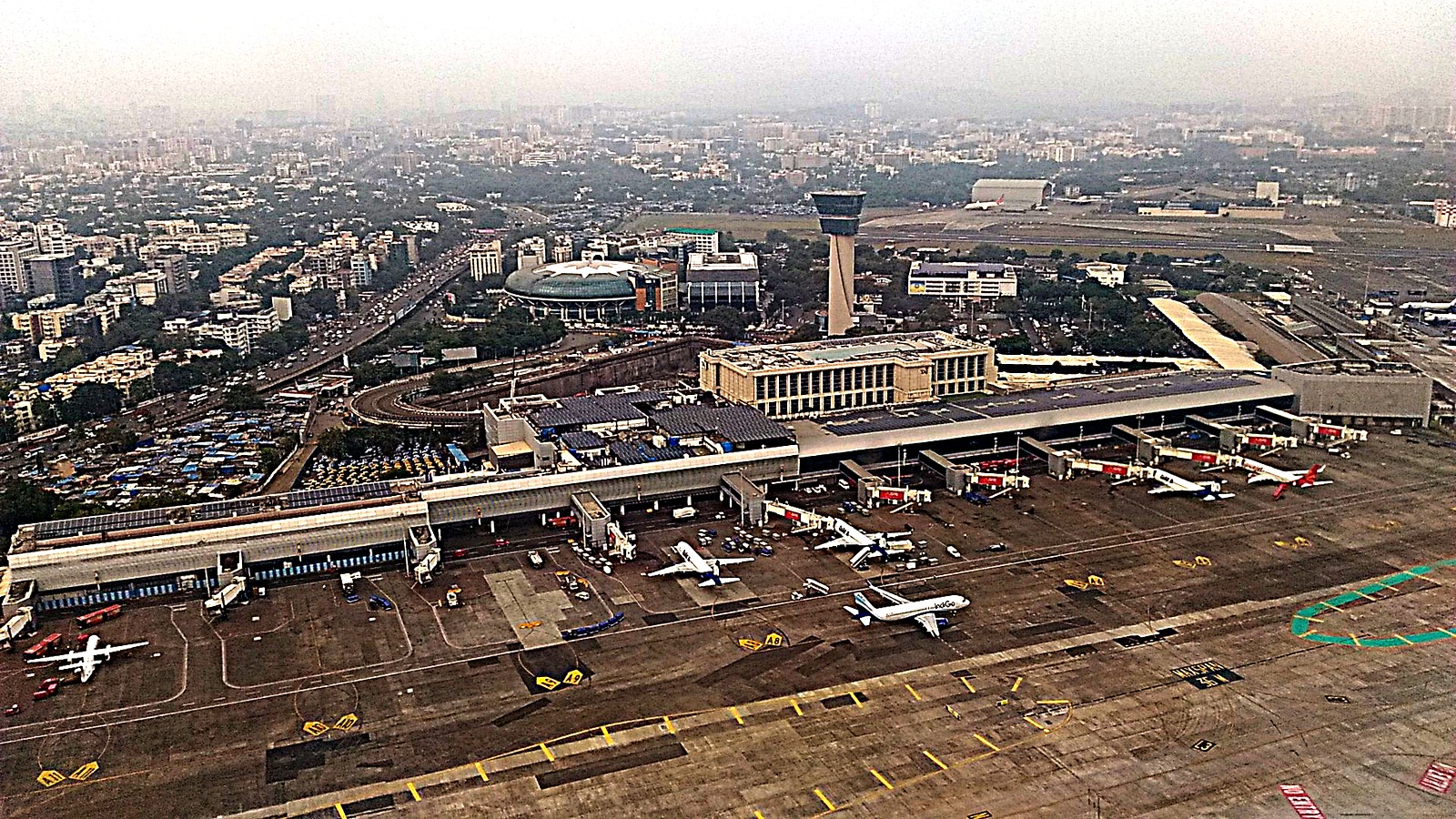 Aerial view of Terminal 1B at Chhatrapati Shivaji Maharaj International Airport in Mumbai, with aircraft at the gates and the city beyond.