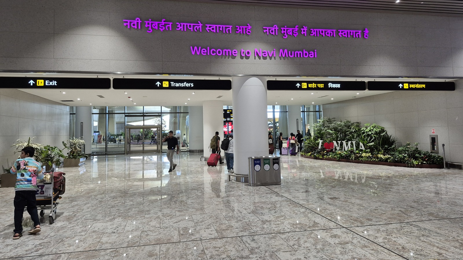 Navi Mumbai International Airport arrivals hall with Welcome to Navi Mumbai signs, Exit and Transfers portals, and an I love NMIA planter