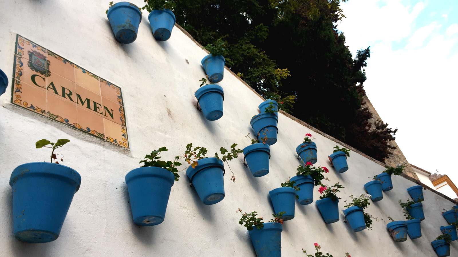 Blue flower pots hang on a whitewashed wall beside a tiled Calle Carmen street sign in Marbella's old town.