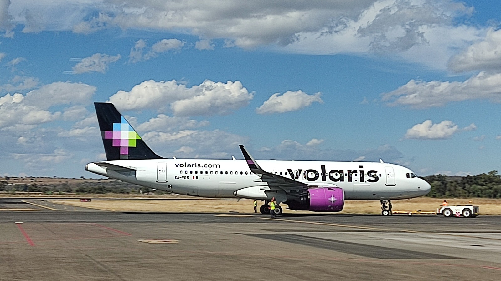 A white Volaris Airbus A320neo with a purple engine being towed on the apron at Morelia airport under a blue sky.