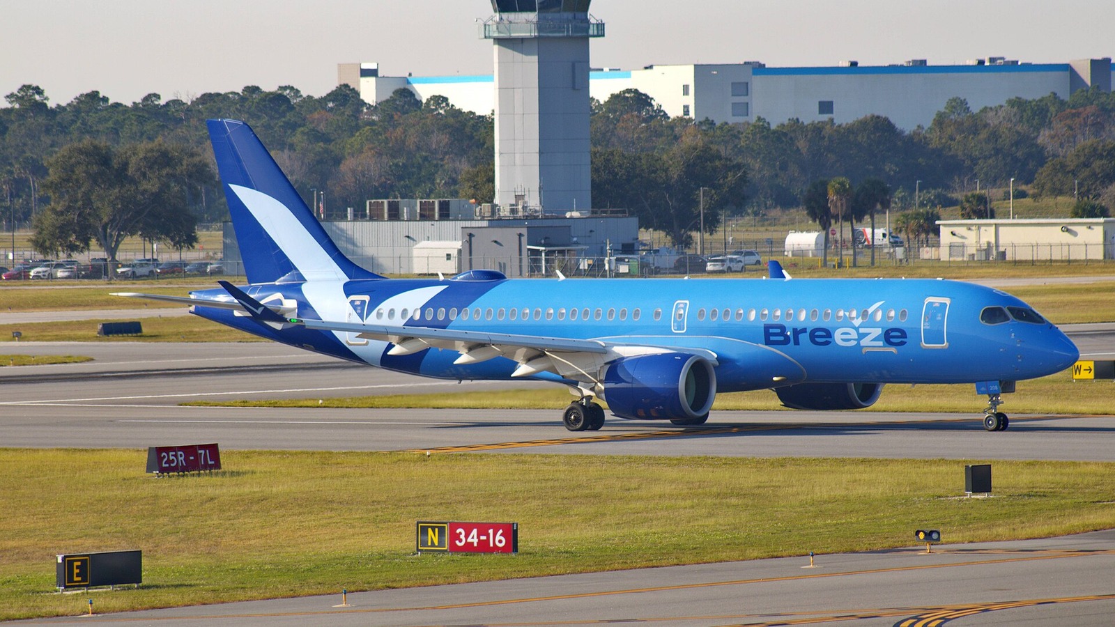 A blue Breeze Airways Airbus A220 taxiing past an airport control tower on a sunny day