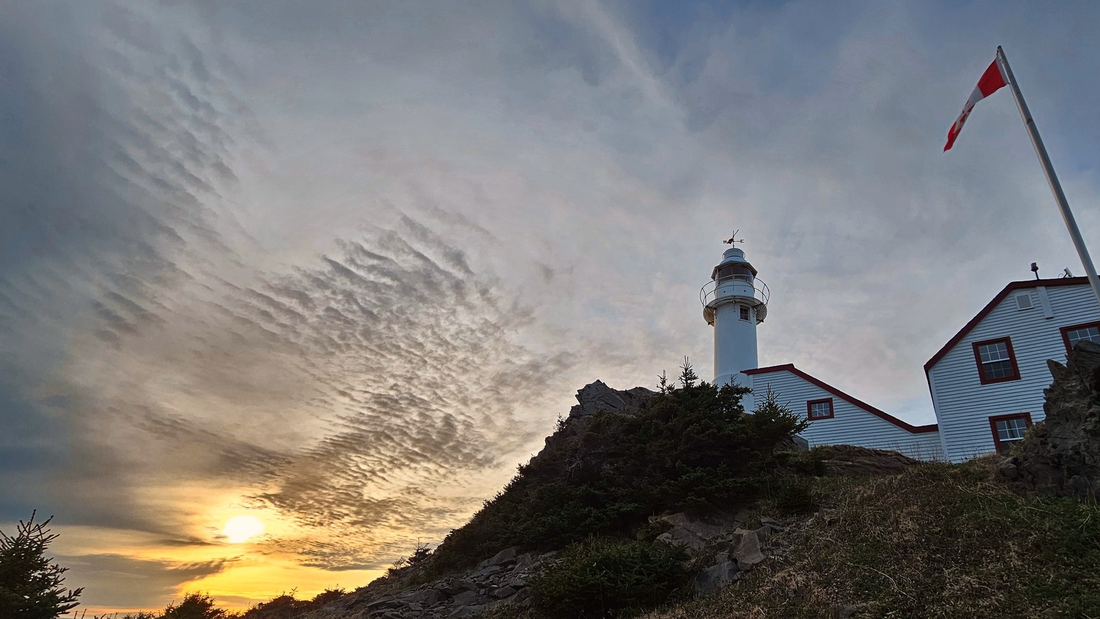 Lobster Cove Head Lighthouse tower and keeper's building on a rocky headland at sunset with a Canadian flag