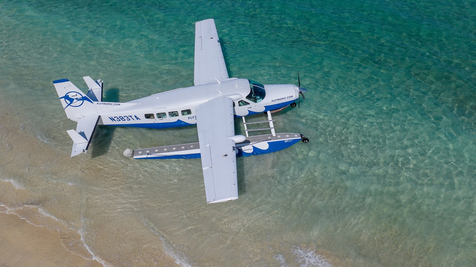 A white and blue Tropic Ocean Airways amphibious seaplane resting in shallow turquoise water near a sandy beach.