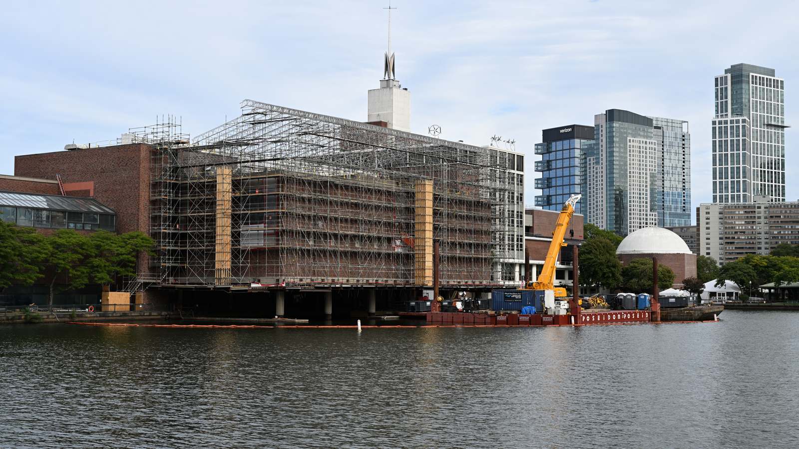 The Museum of Science in Boston seen across the Charles River in September 2024, wrapped in scaffolding with a crane barge alongside during construction.