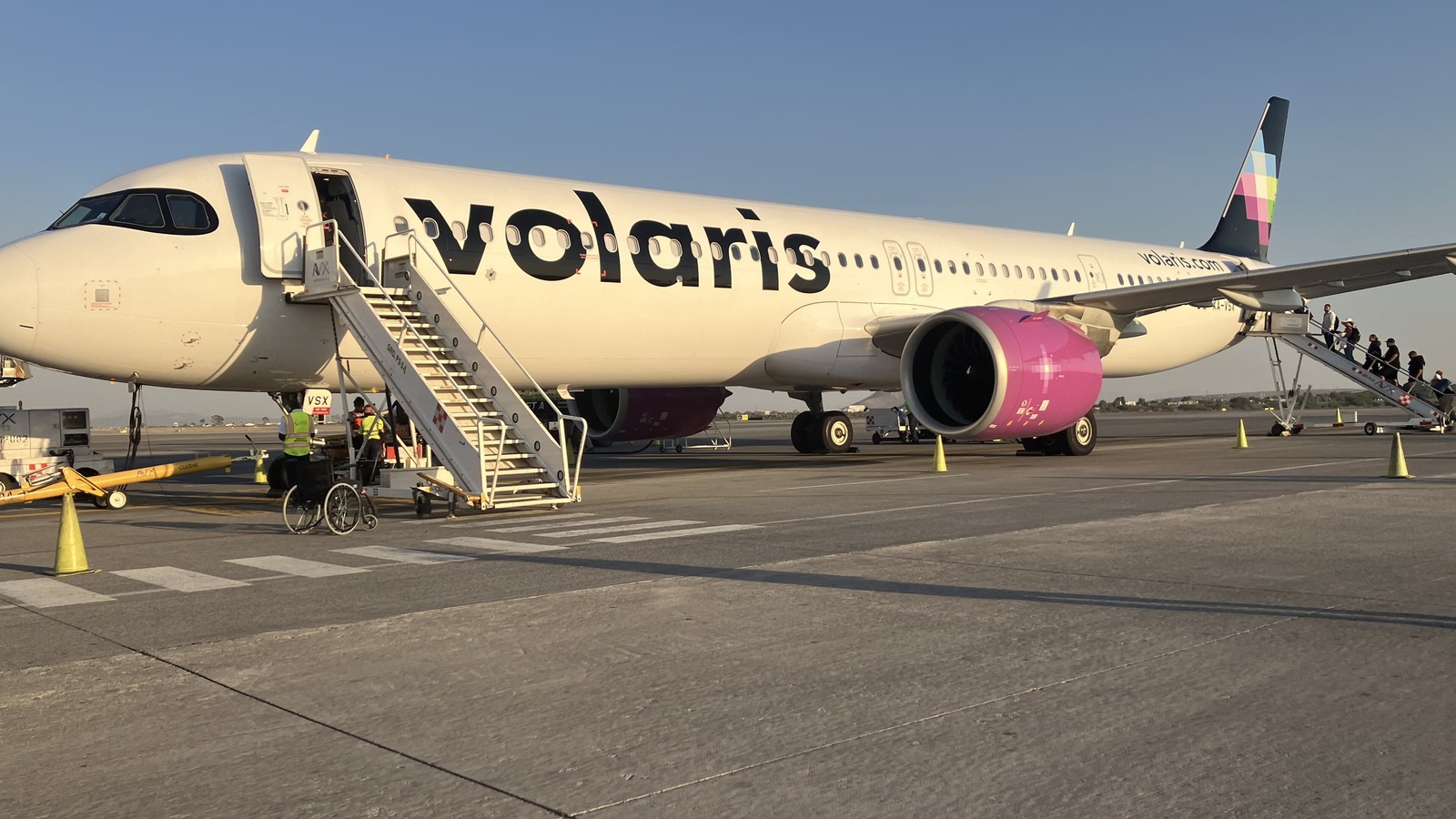 A white Volaris Airbus A321neo with a pink engine and colorful tail parked on an airport apron as passengers board by stairs.