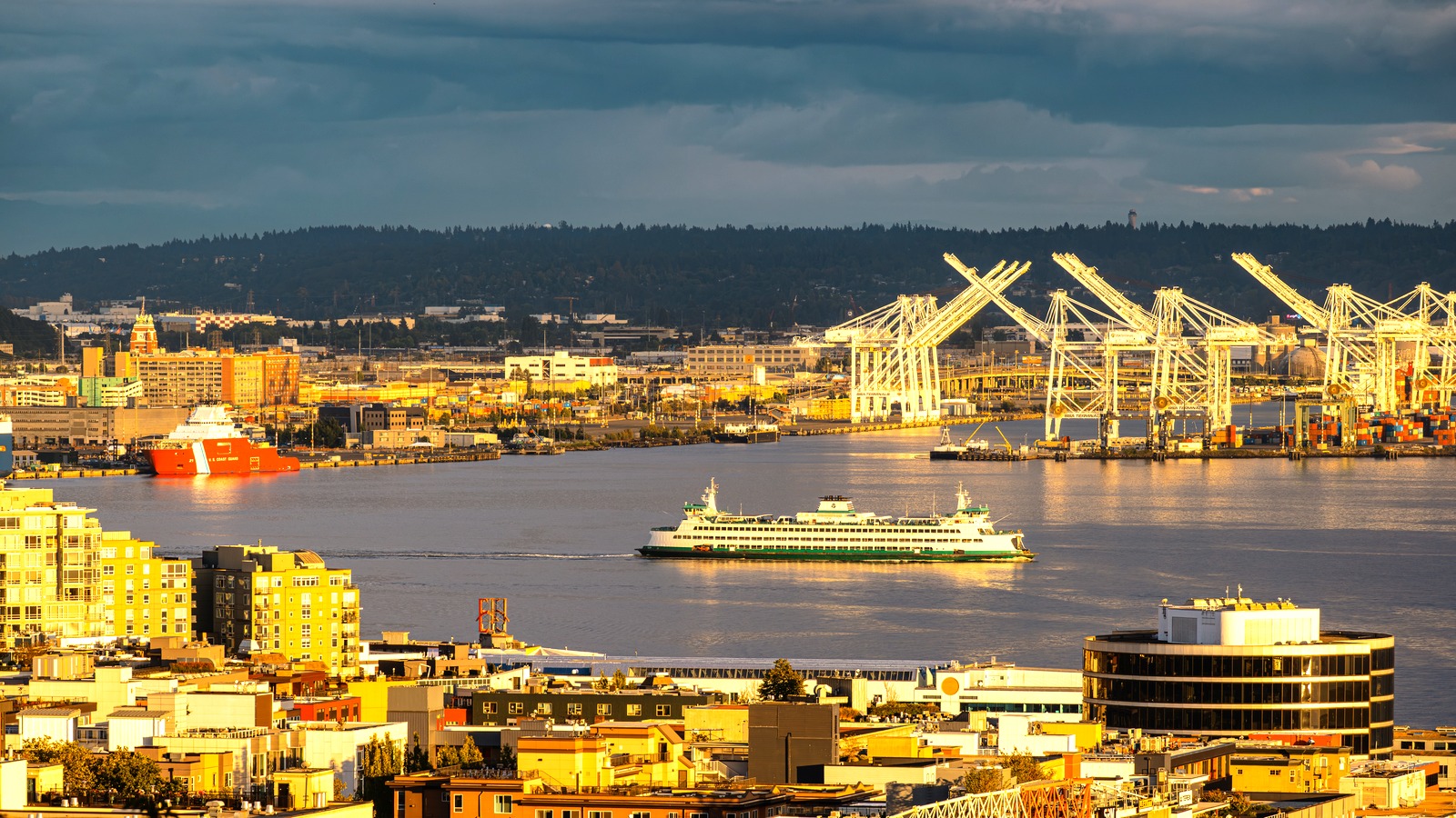 A Washington State Ferries vessel crossing Elliott Bay with Port of Seattle cranes and a U.S. Coast Guard cutter in the background, viewed from Kerry Park in June 2026.