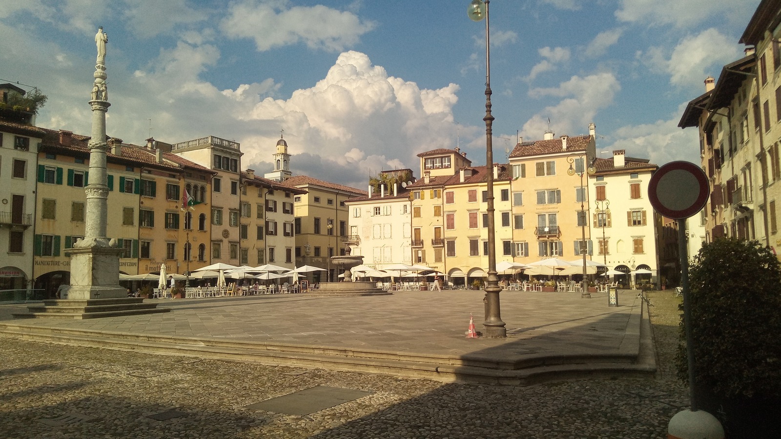 Sunlit Piazza Matteotti in Udine, Italy, with cafe umbrellas and the square visitors cross when city buses thin