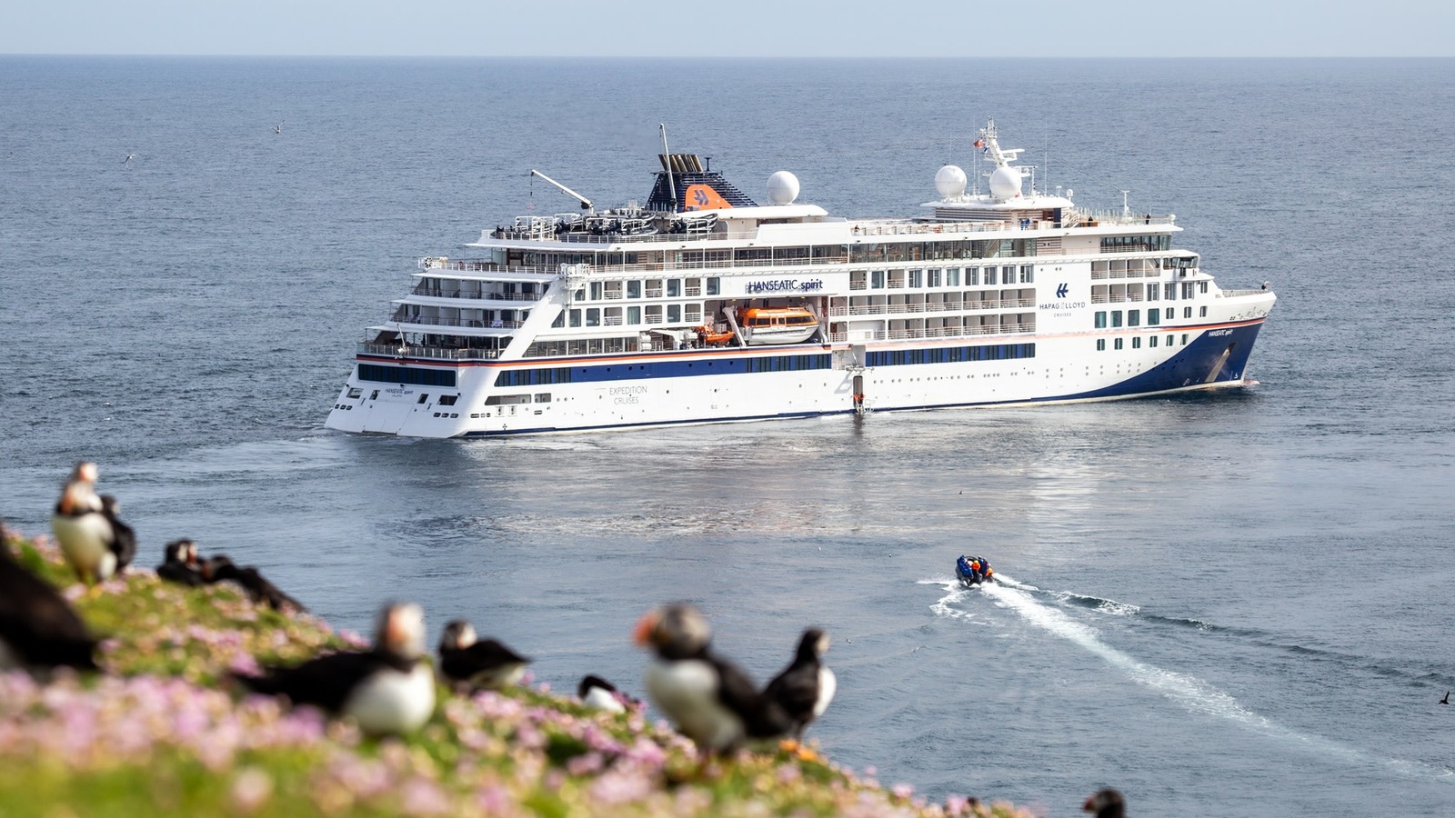 Expedition ship HANSEATIC spirit at sea off a cliff of puffins with a Zodiac heading to shore