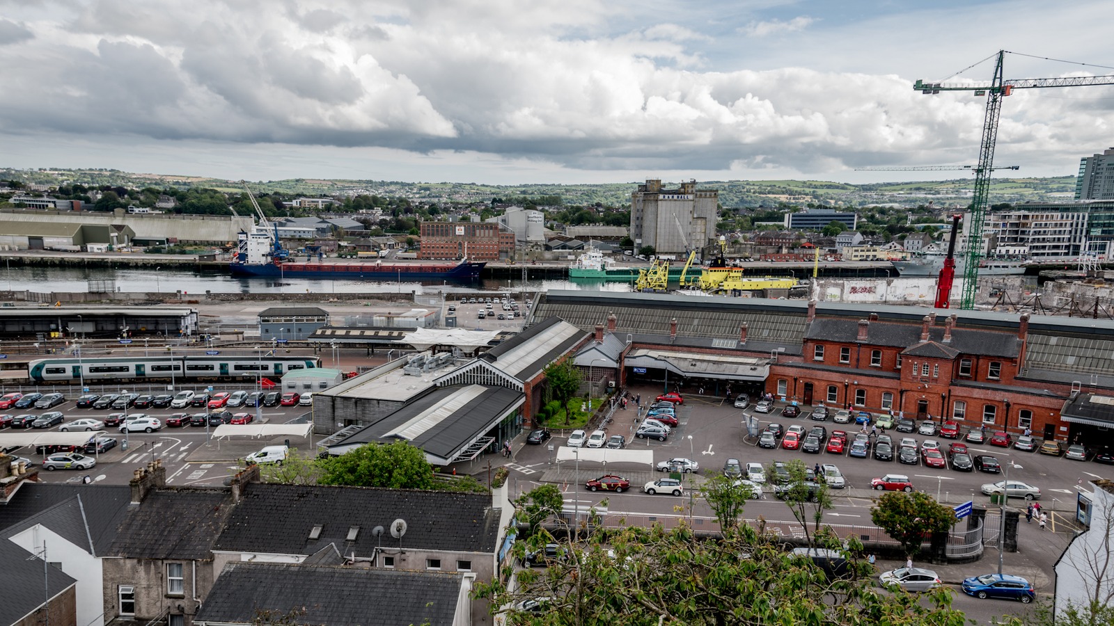 Aerial view of Cork Kent railway station, red-brick sheds, parked cars, and an Irish Rail train beside the River Lee