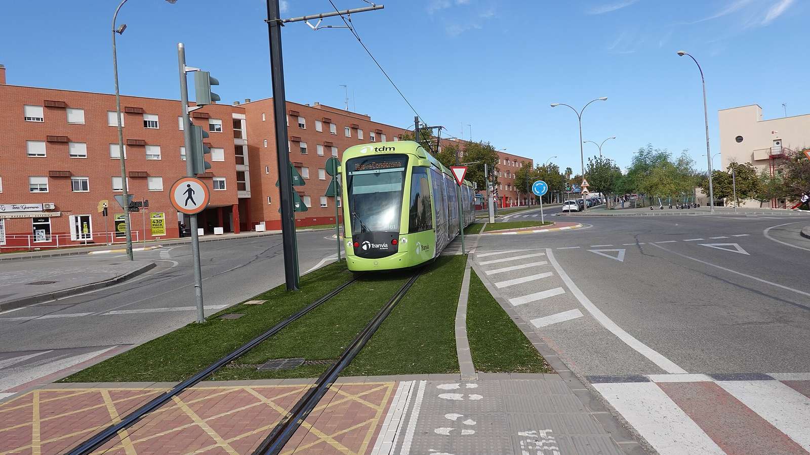 File photo of a green Tranvia de Murcia tram approaching along grass-covered tracks beside a roundabout and red brick apartment blocks under a blue sky