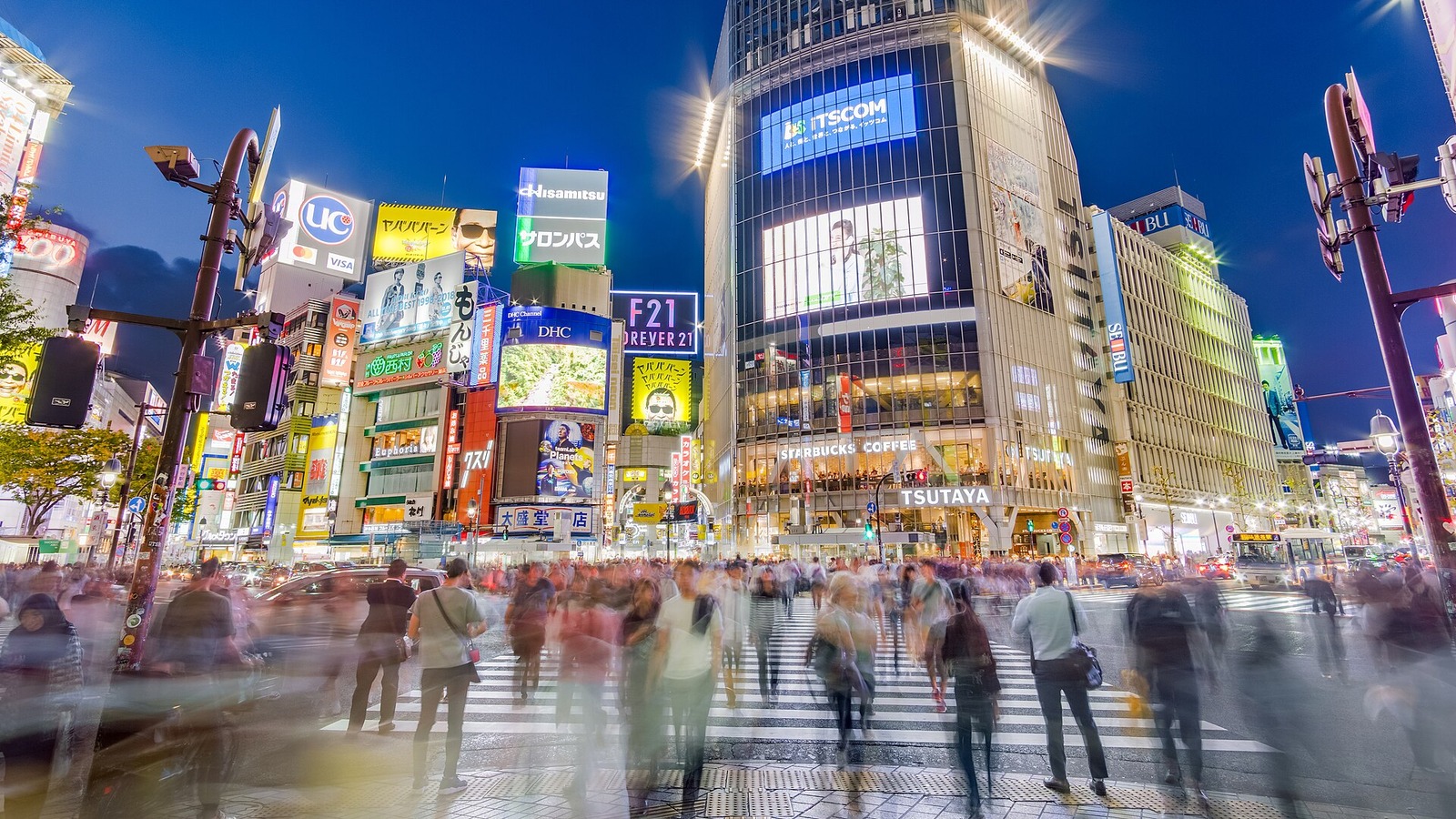 Pedestrians in motion blur cross the Shibuya scramble crossing at dusk beneath lit billboards