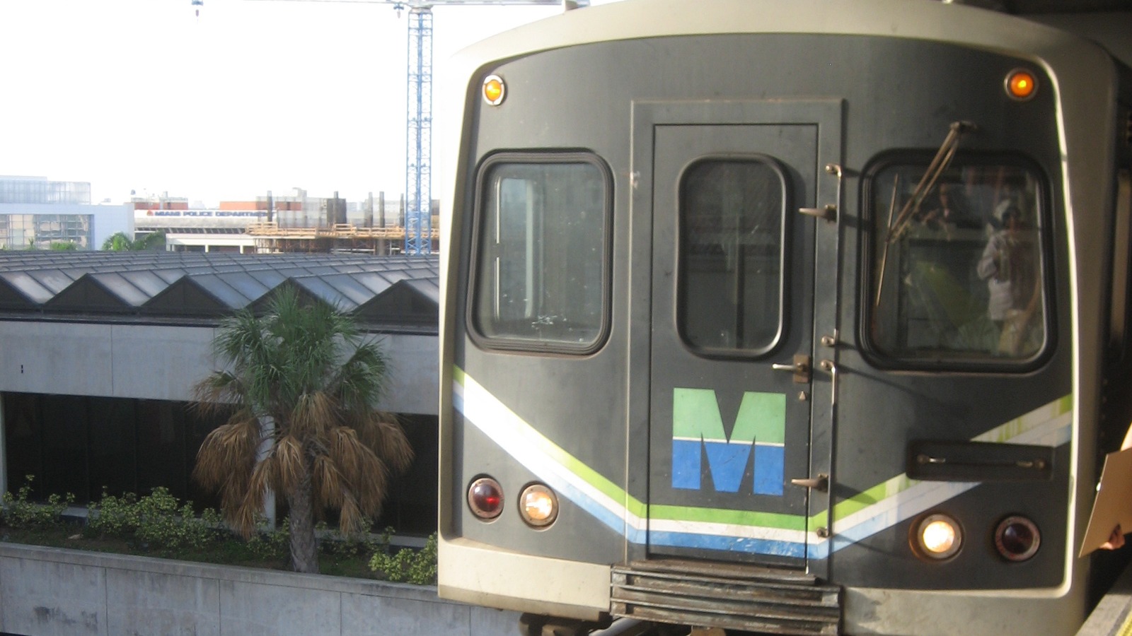 A Miami-Dade Metrorail train with the system M logo arriving at an elevated platform in daylight.