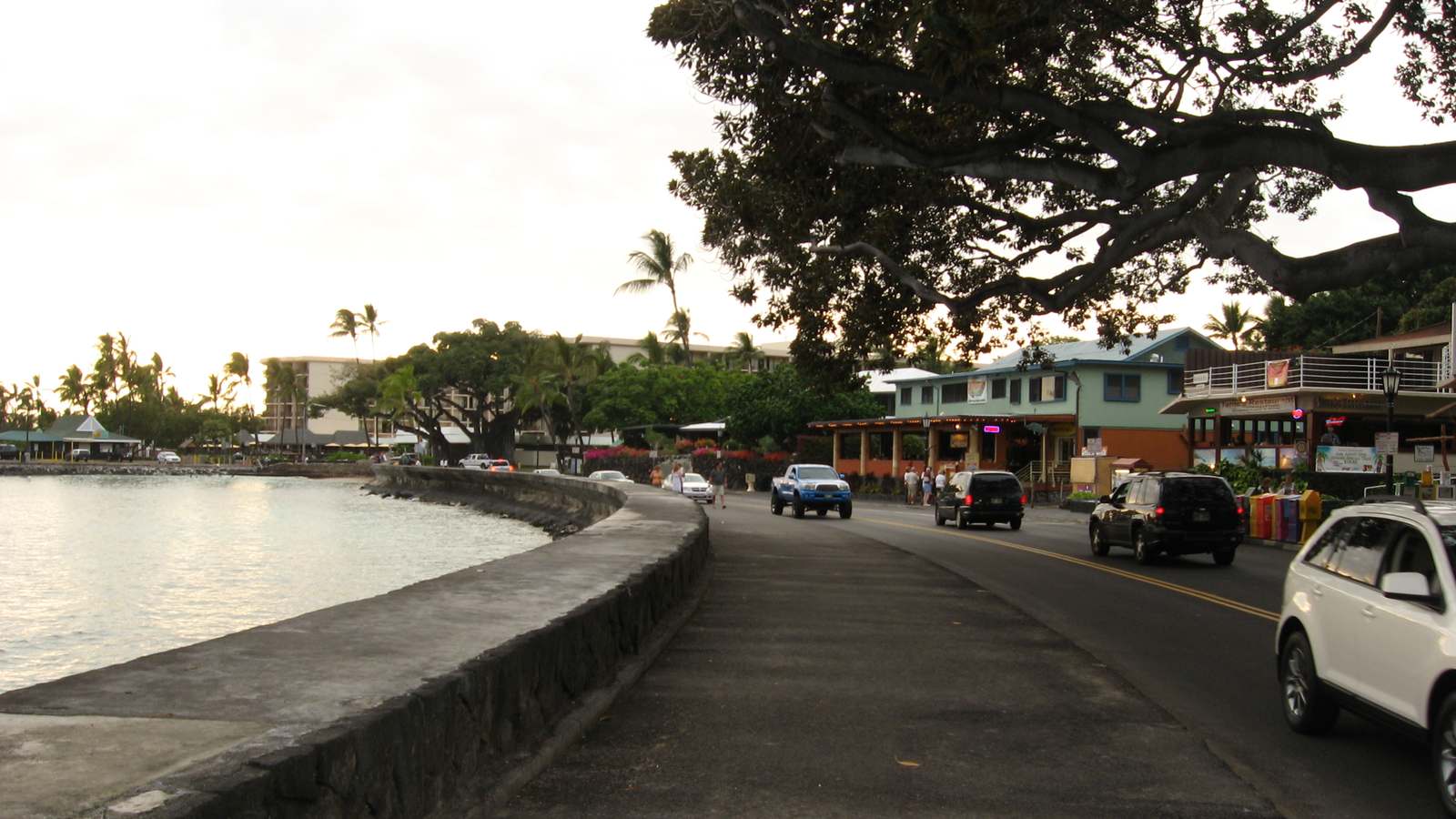 Ali'i Drive curves along the seawall in Kailua-Kona, with shops, banyan trees and palms beside the water.
