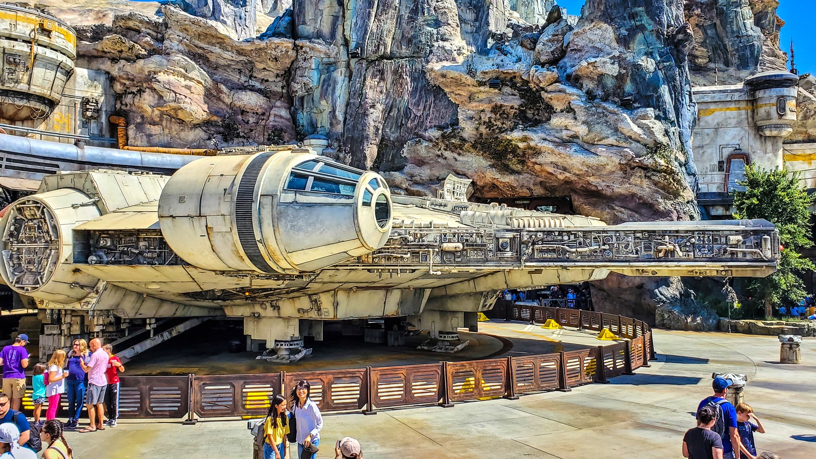 Millennium Falcon replica on the plaza at Star Wars Galaxy's Edge in Disneyland Park, Anaheim, under a clear sky with guests walking in the foreground.