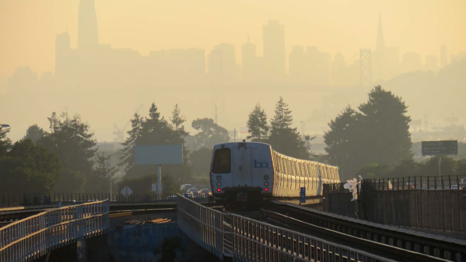 A BART train heads toward San Francisco on an elevated track with the city skyline and Bay Bridge in a hazy golden sky behind it.
