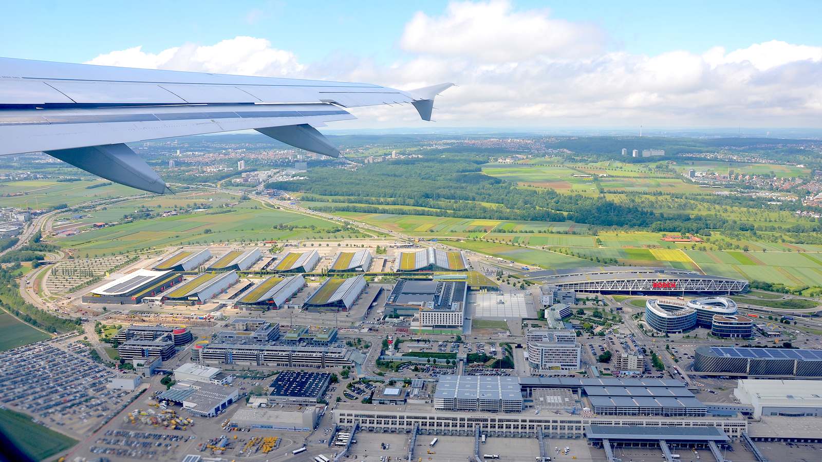 Aerial view of Stuttgart Airport terminals, apron, and the adjacent Stuttgart Trade Fair halls, with an aircraft wing in the foreground