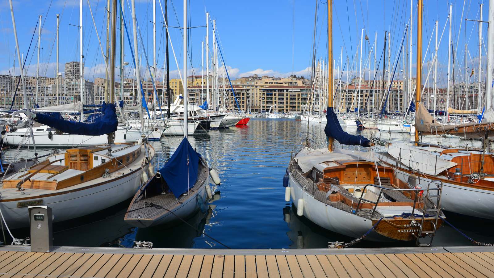Wooden sailboats moored along a boardwalk in Marseille's Vieux-Port under a clear blue sky, with apartment buildings across the water.
