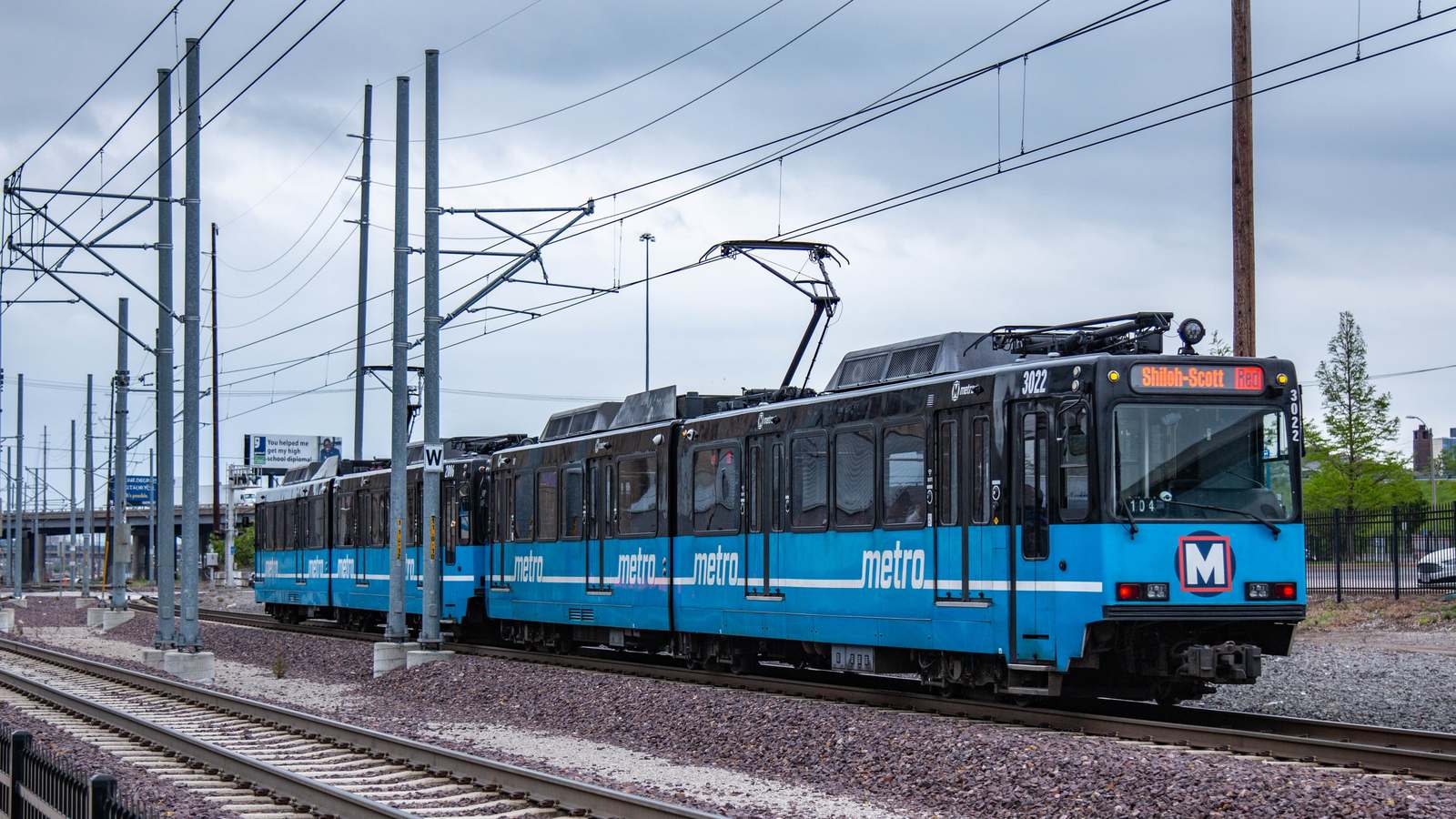 A blue and white MetroLink light rail train with a Shiloh-Scott destination sign runs on tracks under overhead wires on a cloudy day.