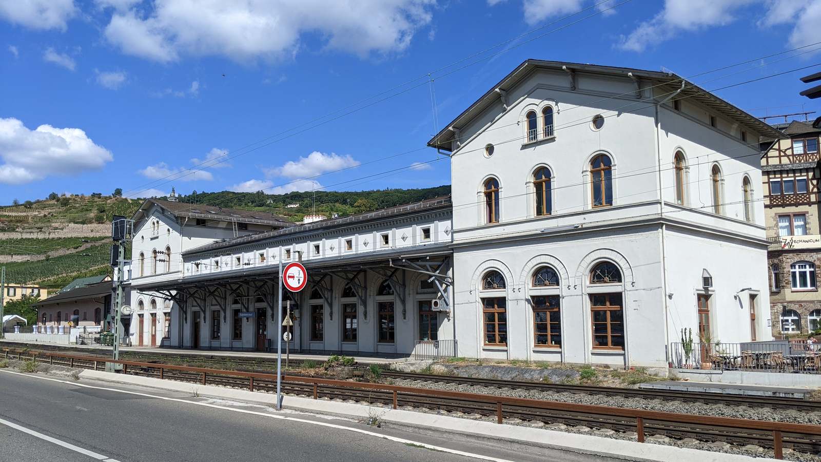 The white two-story station building at Rüdesheim am Rhein beside the tracks with vineyards on the hillside behind