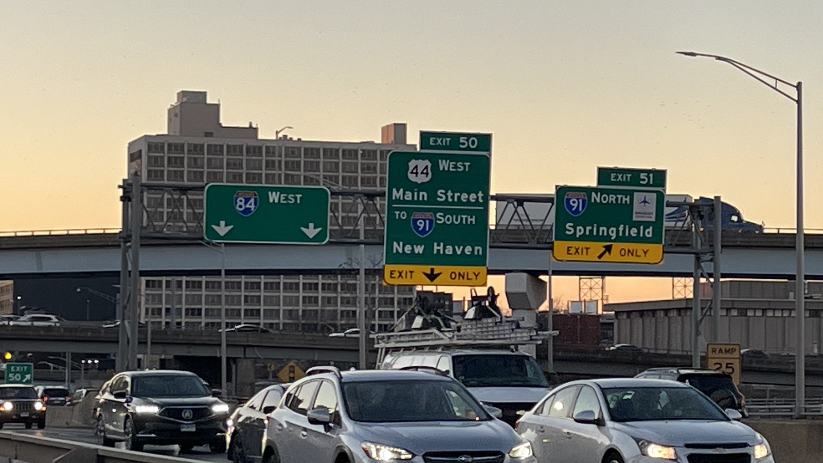Green overhead highway signs for I-84 West, Route 44 West Main Street to I-91 South toward New Haven, and I-91 North toward Springfield at exits 50 and 51 above traffic in Hartford Connecticut at sunset