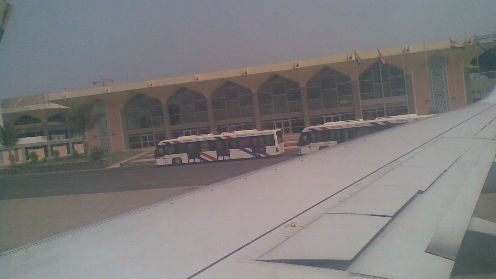 Aden International Airport passenger terminal and apron buses seen from an aircraft wing, with Yemeni flags on the roof