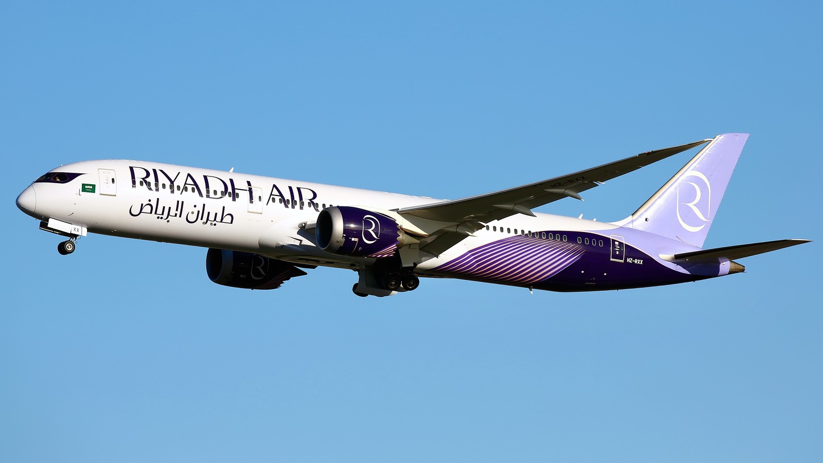 A Riyadh Air Boeing 787-9 in white and purple livery with its landing gear down against a clear blue sky.